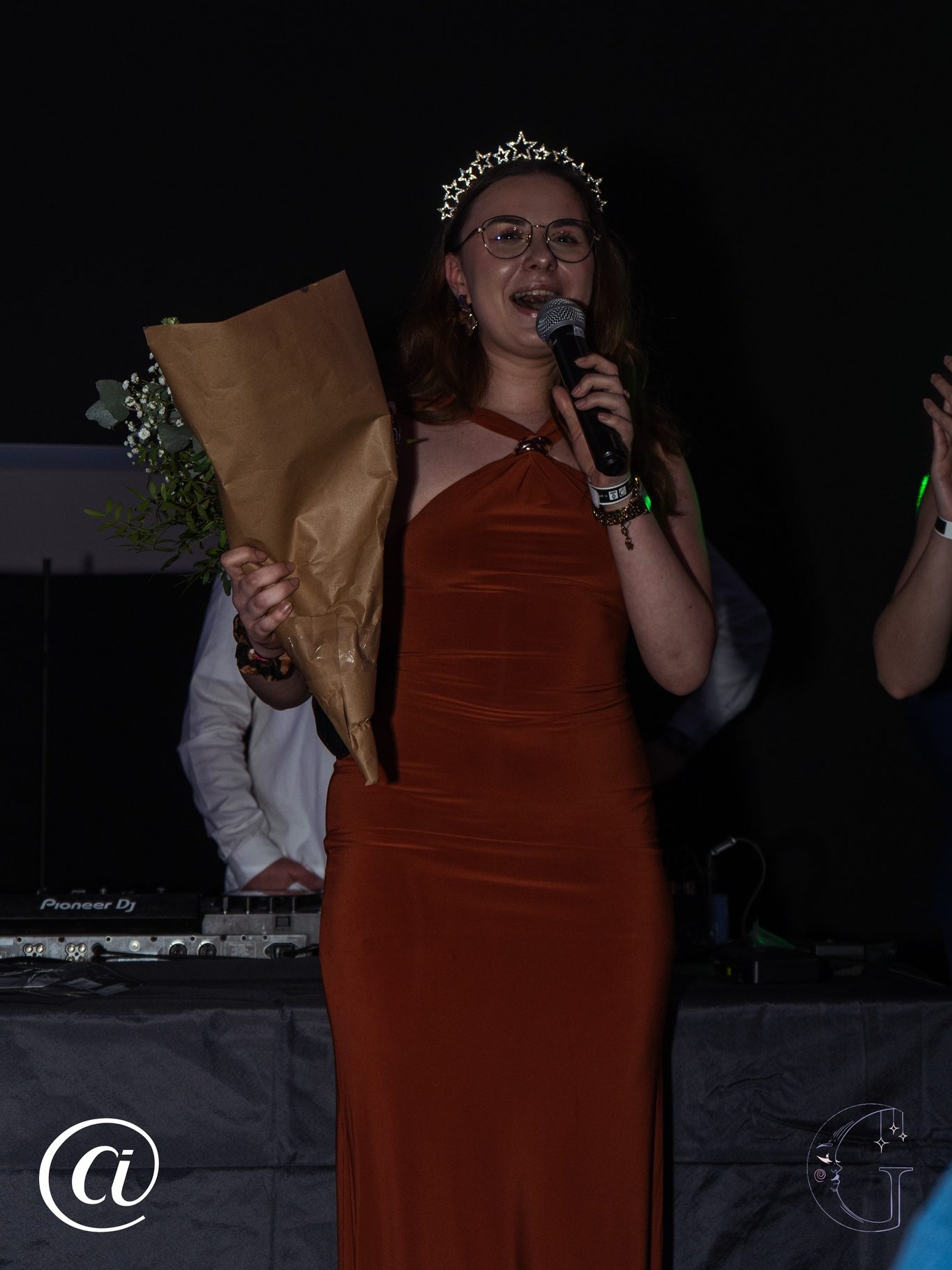 Photo d’une des stars de la Nuit, tenant un bouquet de fleurs dans une main et portant une couronne sur la tête, en train de faire un petit discours au micro.