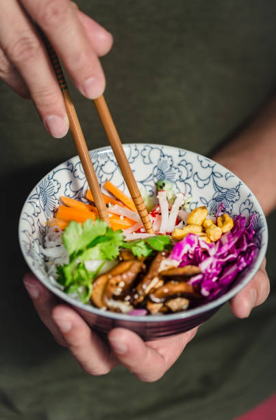 Bol coloré tenu à deux mains, garni de légumes frais; carottes, chou rouge, radis, mais, champignons, noix de cajou et riz. Et on y voit aussi une paire de baguettes en bois tenu dans une mains pour déguster le plat.