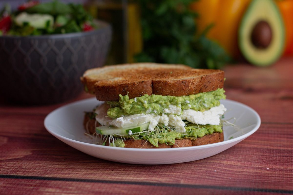 Sandwich garni d’avocat écrasé, fromage frais, tranches de concombre et jeunes pousses, servi dans une assiette blanche sur une table en bois.