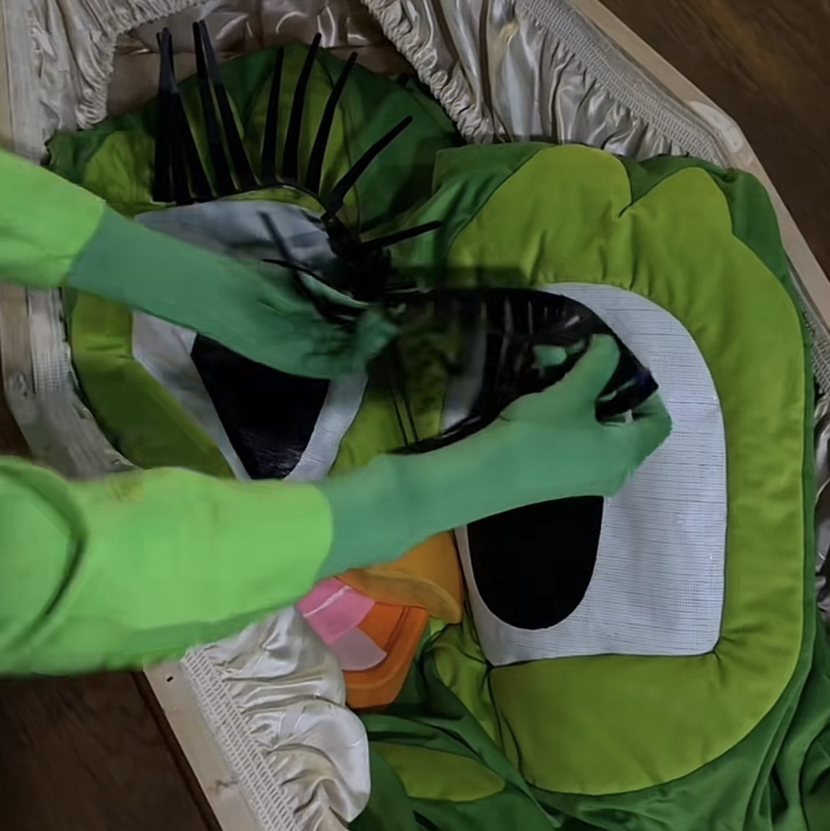 Une personne déguisée en mascotte Duolingo est allongée dans un cercueil ouvert, pendant qu'une autre referme le couvercle. La scène évoque une parodie d'enterrement